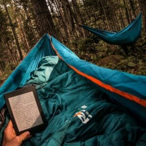 person holding amazon kindle while lying on hammock beside other camper