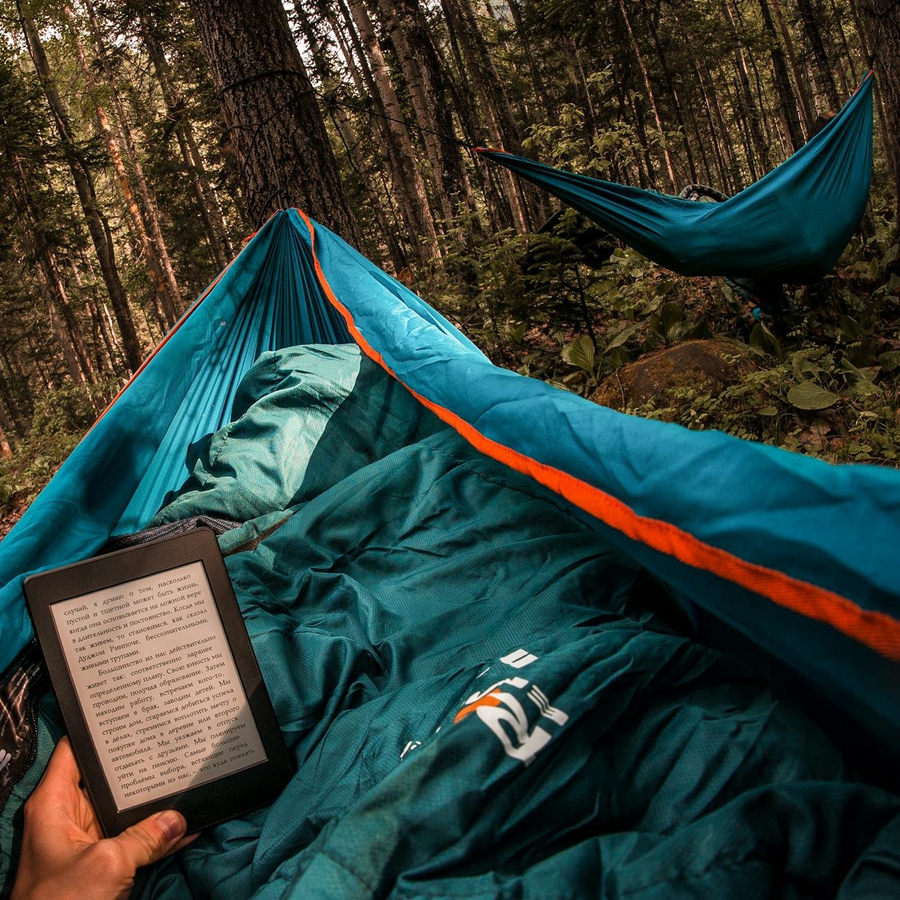 person holding amazon kindle while lying on hammock beside other camper